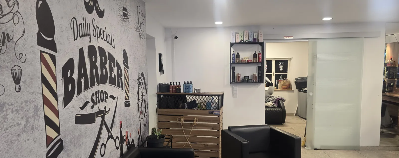 Wartebereich mit schwarzen Ledersesseln, Produktregal und „Barber Shop“-Wandbild